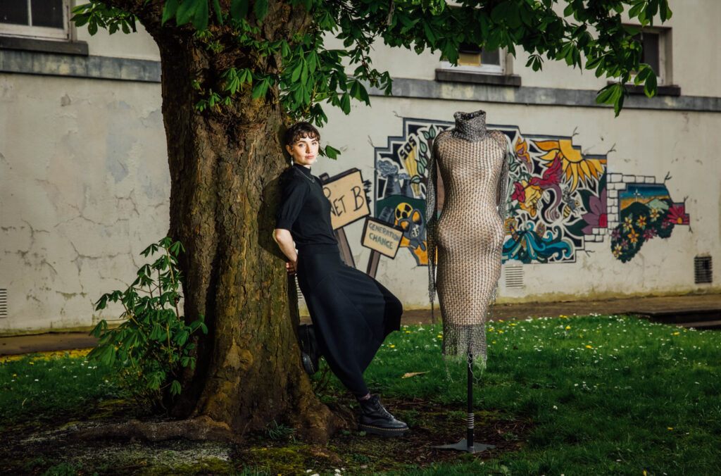 A young woman with short, dark curly hair and a dark outfit leans against a large tree trunk on the left side of the image, looking towards the camera with a slight smile. She is wearing a black short-sleeved top and wide-leg black trousers, along with dark boots. To the right of the tree, a mannequin displays a unique garment made of what appears to be chainmail or metallic mesh, covering the torso and arms. The mannequin stands on a black base. Behind them, there's a light-colored wall with vibrant graffiti featuring abstract shapes, flowers, and patterns in various colors. A small, rustic sign that says "GENERATION CHANGE" is also visible against the wall. The ground is covered with green grass and small white flowers. The overall scene blends natural elements, urban art, and fashion.