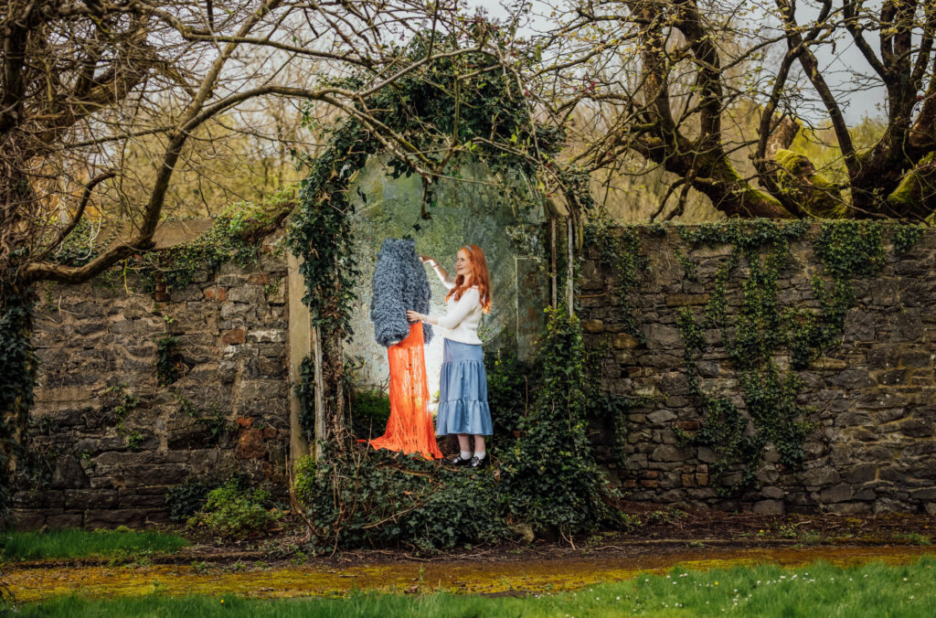 A young woman with long, red hair, wearing a white cardigan and a light blue pleated skirt, stands in front of a stone wall. She is positioned to the right of a mannequin, which is dressed in a vibrant orange skirt and a fluffy dark blue top. The mannequin is placed within a stone archway that is heavily overgrown with ivy and bare branches, creating a rustic, almost fairytale-like setting. The woman is looking towards the mannequin, possibly adjusting the clothing. The ground in the foreground is green grass, and in the background, bare trees and an overcast sky are visible.