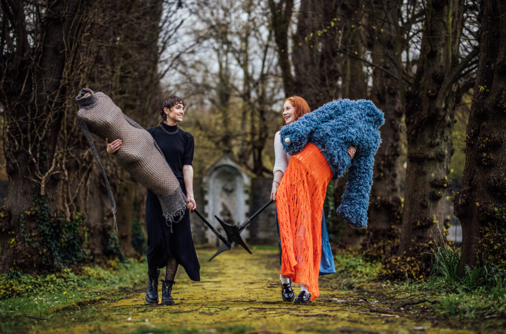 Two young women are walking down a mossy path lined with tall, bare trees, carrying artistic garments or sculptures. The setting appears to be an old, somewhat mystical park or woodland. The woman on the left has short, dark curly hair and is wearing a black long-sleeved dress and dark boots. She is smiling and looking towards the woman on the right, carrying a large, elongated, textured grey fabric sculpture or garment over her shoulder, and a dark, multi-pronged object in her left hand. The woman on the right has long, red hair and is wearing a white long-sleeved top, a vibrant orange skirt, and a light blue undergarment, along with dark shoes. She is also smiling and looking at the woman on the left, carrying a voluminous, fluffy dark blue garment over her arm, and a dark, multi-pronged object similar to the other woman's. In the background, a small stone archway or folly is visible between the trees. The ground is a mix of green grass and patches of yellow-green moss.