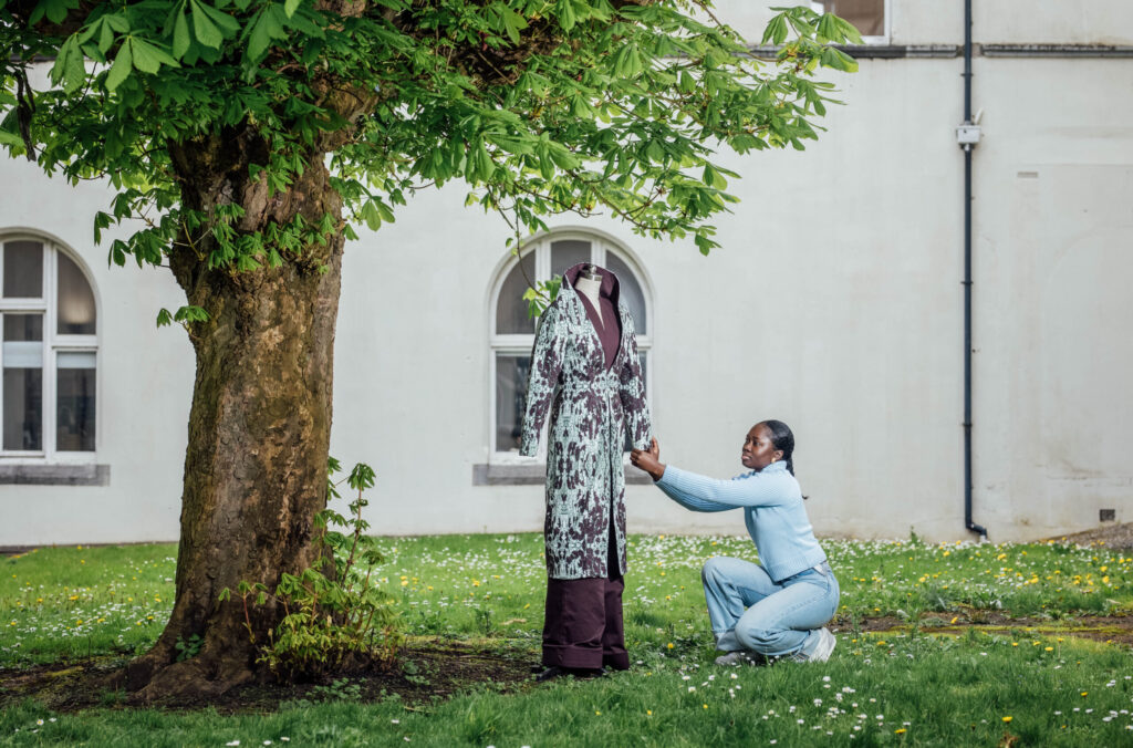 A Black woman with dark hair pulled back, wearing a light blue long-sleeved top and light blue jeans, is kneeling on the grass next to a mannequin. She is adjusting a long, patterned coat on the mannequin, which is dressed in a dark purple garment underneath the coat. The coat has a light green background with darker brown patterns. The scene is outdoors, with a large tree with green leaves on the left, casting some shadow. In the background, there's a light-colored building with arched windows. The ground is covered in green grass with small white flowers.