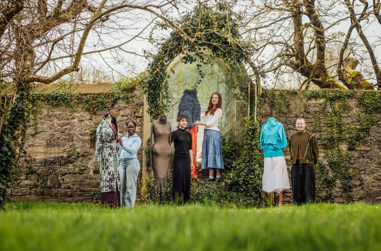 Five people and four mannequins are arranged in a line in front of an old stone wall, heavily covered in ivy and bare tree branches, giving a rustic, artistic backdrop. The ground is covered in vibrant green grass. From left to right: A mannequin wearing a long, patterned coat with shades of green and brown over a dark purple garment. A Black woman with dark hair, wearing a light blue long-sleeved top and light blue jeans, stands next to the mannequin. A mannequin dressed in a textured, light brown or grey, form-fitting garment. A young woman with short, dark curly hair, wearing a black long-sleeved dress, stands next to the textured mannequin. A young woman with long, red hair, wearing a white cardigan and a light blue pleated skirt, stands next to a mannequin dressed in an orange skirt and a fluffy dark blue top. This mannequin is placed within a stone archway overgrown with ivy. The red-haired woman appears to be interacting with the mannequin's top. A mannequin wearing a light blue top and a white, sheer skirt with a rough hem. A person with short, light brown hair, wearing a dark green or brown jacket and dark trousers, stands next to the last mannequin. The individuals are looking at the camera or towards the art pieces, and the overall scene appears to be an outdoor fashion or art exhibition.