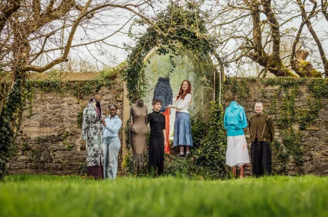 Five people and four mannequins are arranged in a line in front of an old stone wall, heavily covered in ivy and bare tree branches, giving a rustic, artistic backdrop. The ground is covered in vibrant green grass. From left to right: A mannequin wearing a long, patterned coat with shades of green and brown over a dark purple garment. A Black woman with dark hair, wearing a light blue long-sleeved top and light blue jeans, stands next to the mannequin. A mannequin dressed in a textured, light brown or grey, form-fitting garment. A young woman with short, dark curly hair, wearing a black long-sleeved dress, stands next to the textured mannequin. A young woman with long, red hair, wearing a white cardigan and a light blue pleated skirt, stands next to a mannequin dressed in an orange skirt and a fluffy dark blue top. This mannequin is placed within a stone archway overgrown with ivy. The red-haired woman appears to be interacting with the mannequin's top. A mannequin wearing a light blue top and a white, sheer skirt with a rough hem. A person with short, light brown hair, wearing a dark green or brown jacket and dark trousers, stands next to the last mannequin. The individuals are looking at the camera or towards the art pieces, and the overall scene appears to be an outdoor fashion or art exhibition.