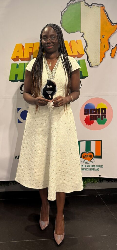 Sherifat Ukachukwu, Clinical Skills Learning Coordinator at Technological University of the Shannon (TUS), poses with the Outstanding African Healthcare Leader Award at the African Health Summit Ireland conference in the Crowne Plaza Hotel, Blanchardstown, Dublin. She is wearing a cream dress and holding the award in front of an event backdrop featuring an outline of Africa and summit branding.