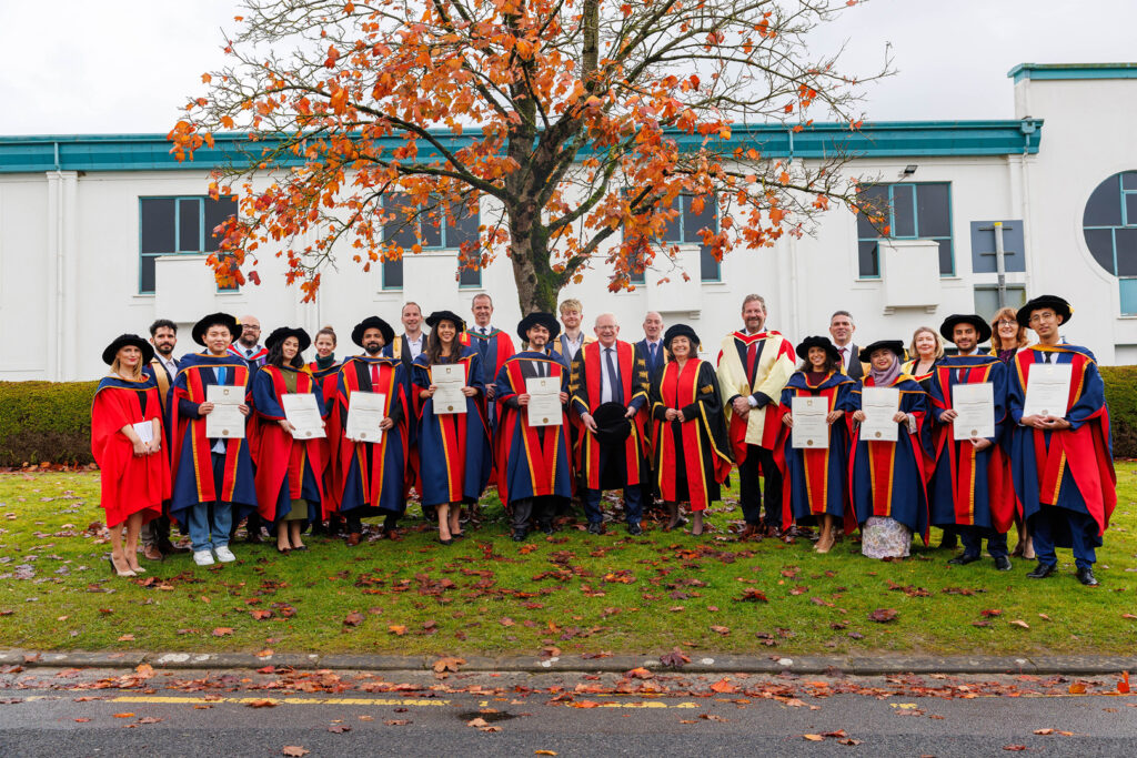 TUS PhD candidates stand on the grass underneath a horse chestnut tree holding up their graduation parchments and smiling, wearing red and navy ceremonial graduation attire.