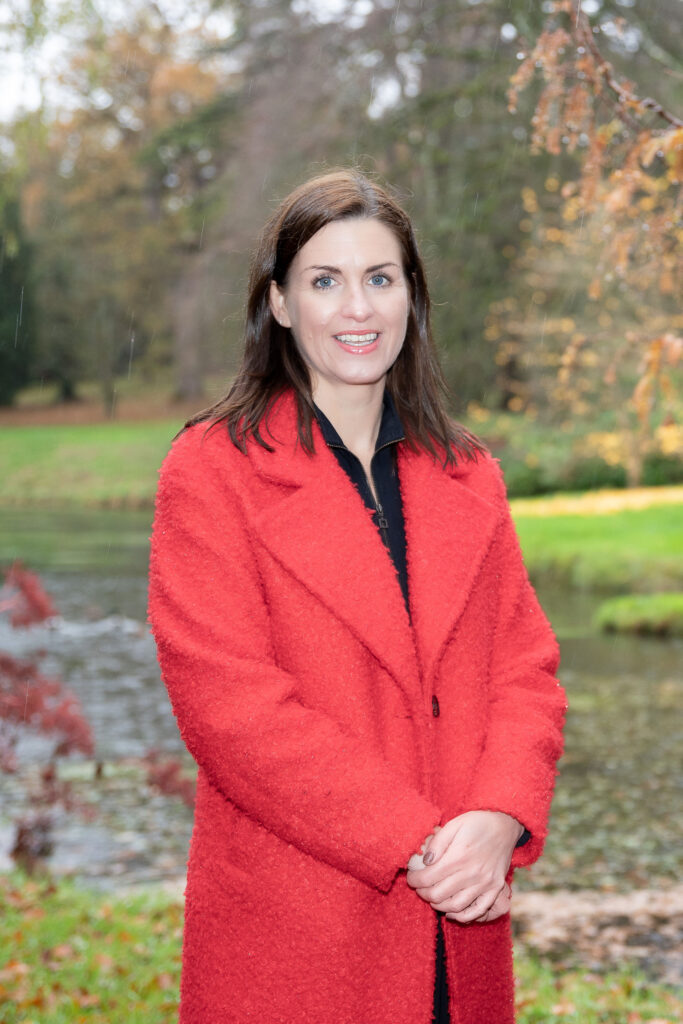 A woman with long, dark brown hair and blue eyes smiles at the camera. She is wearing a textured, bright red coat over a black top, with her hands clasped in front of her. She is standing outdoors in what appears to be a park or garden setting, with a body of water behind her and trees with autumn foliage. The background is slightly blurred, giving emphasis to the woman.