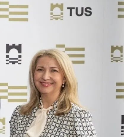 A smiling woman with long, blonde hair and light makeup looks directly at the camera. She is wearing a patterned blouse with dark blue geometric shapes on a white background and a light-colored bow tied at the neck. She also wears small pearl earrings. The background is a white wall decorated with multiple 