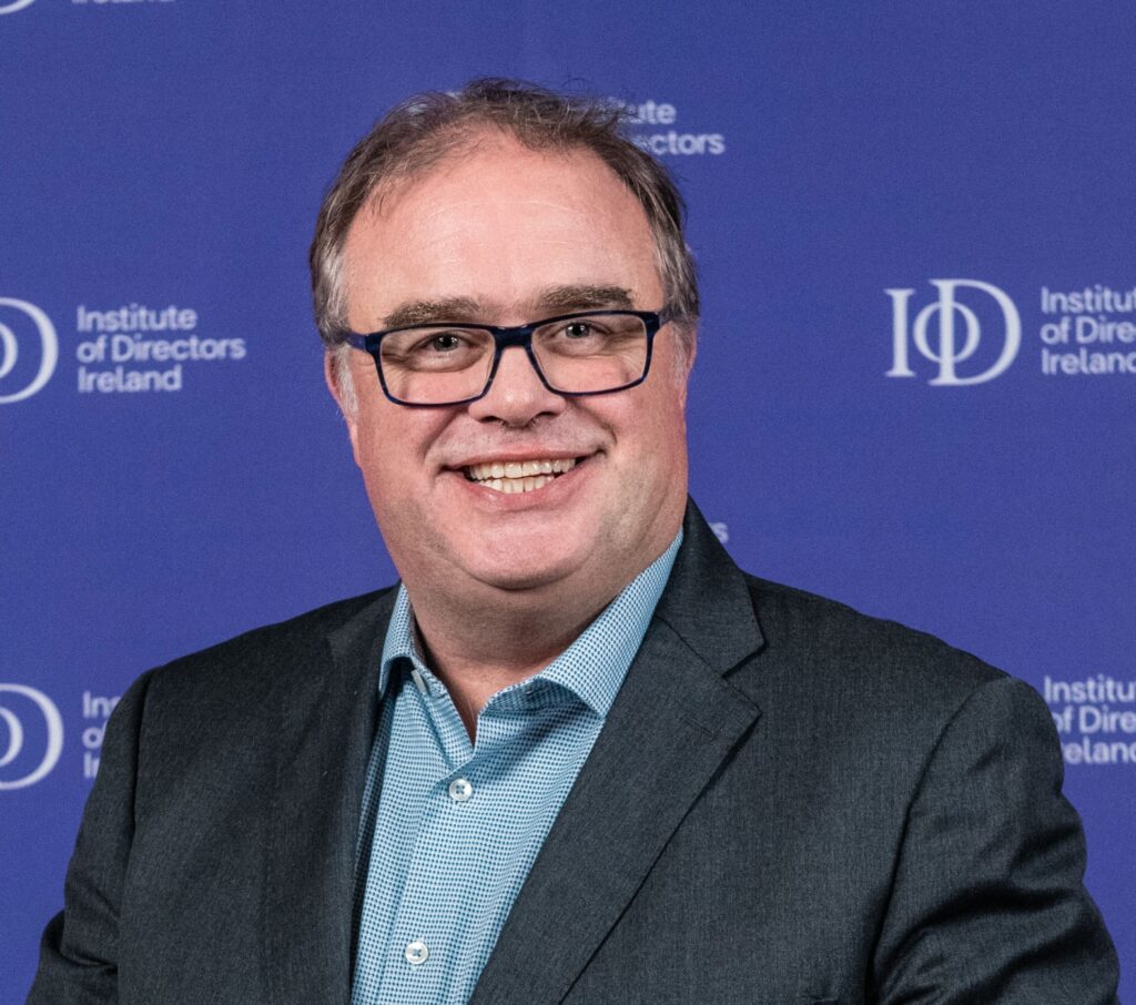 A man with light brown hair, wearing glasses, smiles broadly at the camera. He is wearing a dark grey suit jacket over a blue checkered collared shirt. The background is a vibrant blue wall with the "Institute of Directors Ireland" logo partially visible in white several times.