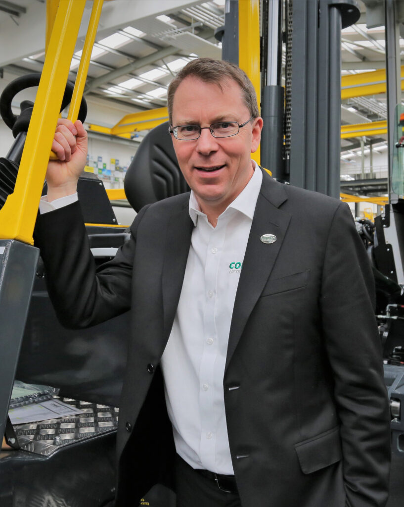 A man with short brown hair and glasses, wearing a black suit jacket over a white collared shirt, smiles faintly at the camera. He is holding onto a yellow bar of what appears to be a large industrial machine, possibly a forklift, in a factory or warehouse setting. A green logo is visible on his shirt. The background shows parts of the industrial machinery and the interior of a facility.