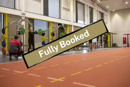 An indoor gym or training facility with an orange track and various weightlifting equipment. In the foreground, a large, angled banner in a khaki color with a dark border reads 