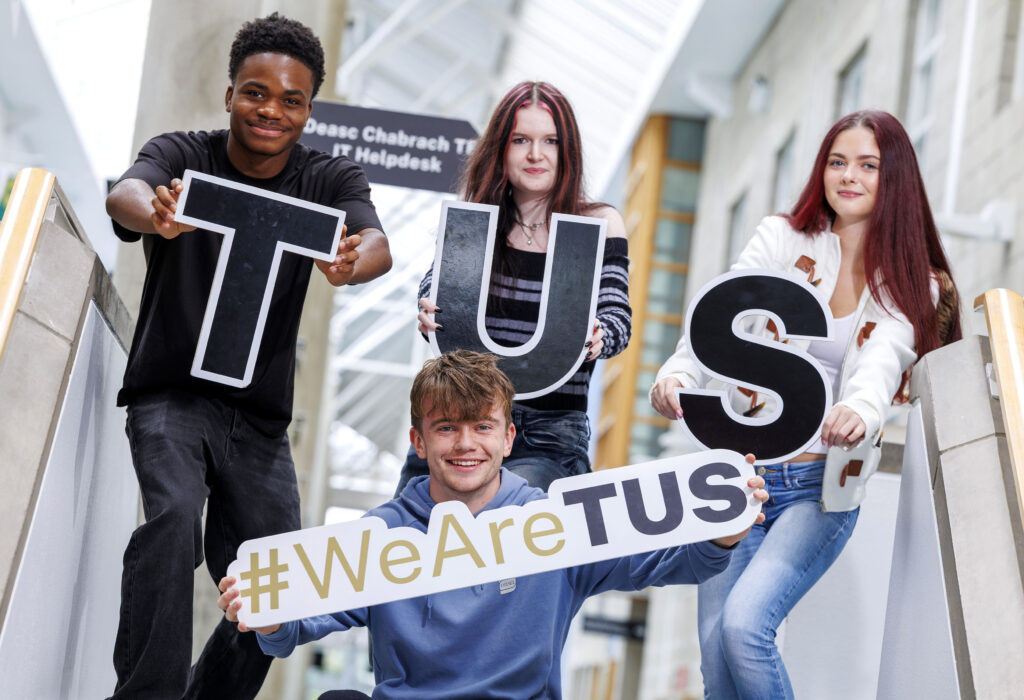 Four young adults are posing on a staircase, holding up letters that spell "TUS" and a sign that says "#WeAreTUS." From left to right: A young Black man with a dark t-shirt and black pants smiles at the camera, holding a large black letter "T" with a white border. Behind him, a young woman with dark hair and red streaks, wearing a striped long-sleeved top and jeans, holds up a large black letter "U" with a white border. In front and center, a young white man with light brown, curly hair, wearing a blue hoodie, kneels and smiles while holding a white sign that reads "#WeAreTUS" in gold and black text. On the far right, a young white woman with long dark red hair, wearing a white jacket over a white crop top and light-wash jeans, holds up a large black letter "S" with a white border. The background shows a bright, modern interior space with stairs and a blurred sign that appears to say "Helpdesk."
