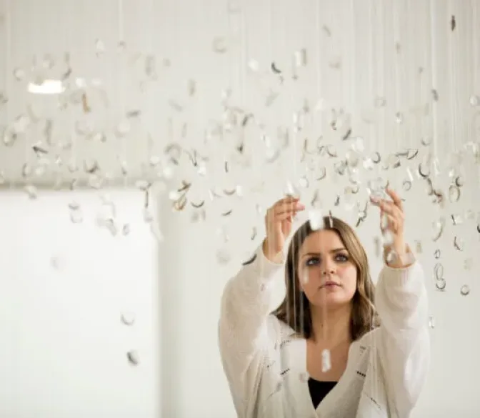 A young woman with light brown hair and a serious expression is standing, looking up at an art installation composed of numerous small, light-colored, possibly translucent or reflective objects suspended from the ceiling by thin wires. She is wearing a white cardigan over a dark top, with her hands raised as if interacting with or observing the suspended elements. The background is a bright, plain white wall, which emphasizes the delicate nature of the artwork.