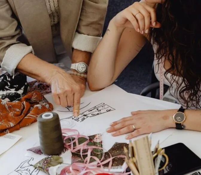 A close-up shot of two people collaborating on a fashion design. An older person, wearing a beige jacket and a watch, points at a drawing on a white sheet of paper with their right hand. The drawing appears to be a fashion sketch with details. Next to them, a younger person with long, dark, curly hair and a white tank top has their left hand on the paper, wearing a watch and rings. A pink measuring tape hangs around their neck. The table is covered with various items including swatches of patterned fabric, spools of thread (one prominent dark green spool), ribbon, scissors, and other design tools. The focus is on their hands and the design process.