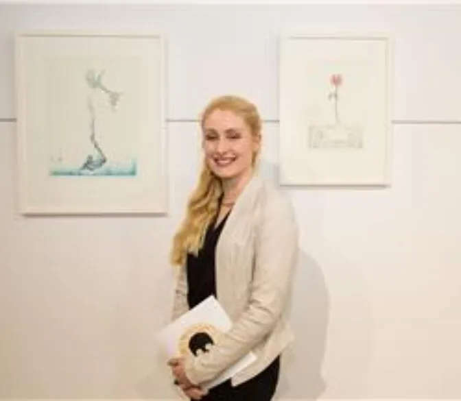 A smiling woman with long, blonde, wavy hair stands in an art gallery, holding a white booklet with a dark logo. She is wearing a cream-colored blazer over a black top. Behind her, two framed artworks are displayed on a white wall. The artwork on the left appears to be a light blue sketch of a figure, while the one on the right is a delicate drawing of a single red flower.
