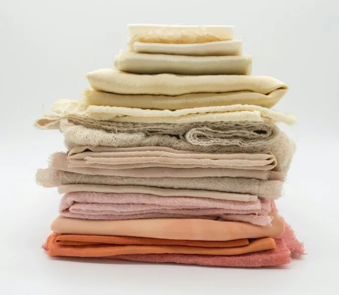A neatly stacked pile of fabric swatches or textiles, ranging in color from white and cream at the top to various shades of beige, light pink, peach, and coral at the bottom. The fabrics appear to be natural materials, possibly linen or cotton, with some having visible textures or slight fraying at the edges. The stack is centered against a plain white background.