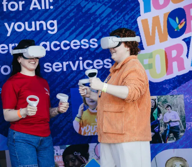 Two young people are smiling and interacting while wearing white virtual reality headsets and holding controllers. The person on the left wears a red t-shirt and denim shorts, with visible tattoos on their arm. The person on the right has reddish-brown curly hair and is wearing an orange textured jacket over a white top. They are standing in front of a vibrant blue backdrop covered with text and images related to 