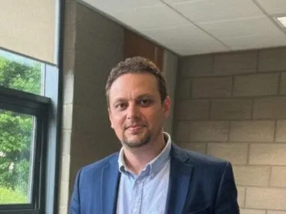 A man with short brown hair and a light goatee smiles faintly while looking directly at the camera. He is wearing a dark blue blazer over a light blue collared shirt with the top button undone. He is indoors, with a window visible on the left side of the frame and a light-colored brick wall behind him on the right. The ceiling has acoustic tiles.