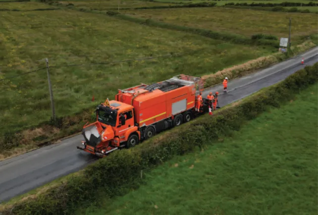 An aerial view of an orange road maintenance truck parked on a narrow paved road, surrounded by lush green fields. The truck is large and appears to be involved in road resurfacing, as part of the road behind it looks freshly paved and darker than the older section. Several construction workers in orange high-visibility clothing are gathered around the rear of the truck, appearing to oversee the work. Traffic cones are placed on the road near the workers. Telephone poles with wires run along the side of the road, and a signpost is visible in the distance.