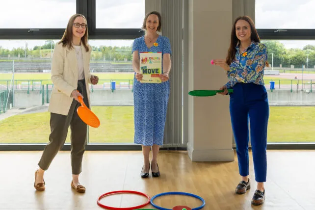 hree women stand in a bright, modern room with large windows overlooking a sports stadium or track. From left to right: A woman with dark hair and glasses, wearing a light-colored patterned blazer over a white top, dark olive trousers, and brown flats, smiles at the camera while holding an orange paddle. A woman with dark hair, wearing a blue floral dress and black heels, smiles at the camera while holding a book or sign that says 
