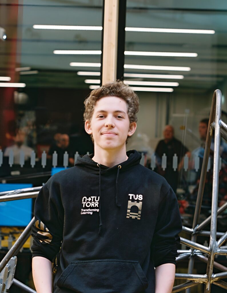 A young man with light brown, curly hair stands facing the camera, smiling slightly. He is wearing a black hoodie with a white logo that says "NTNU TORR Transforming Learning" on the left side of his chest and a gold logo that says "TUS" on the right. His hands are by his side, and he is standing indoors in front of a large glass windows that reflect indoor lighting and faint figures. Behind him is a metal structure.