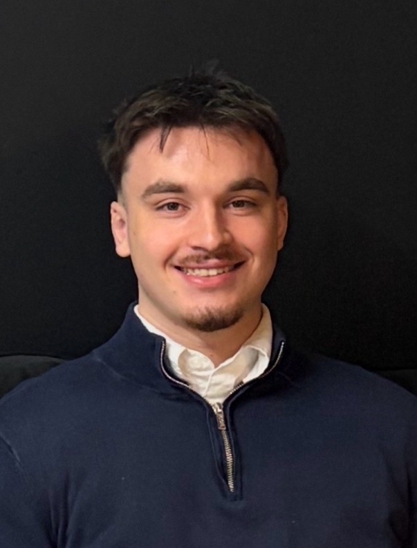 A young man with short dark hair and a light goatee smiles broadly while looking directly at the camera. He is wearing a navy blue quarter-zip sweater over a white collared shirt. The background is a plain dark surface, possibly a wall.