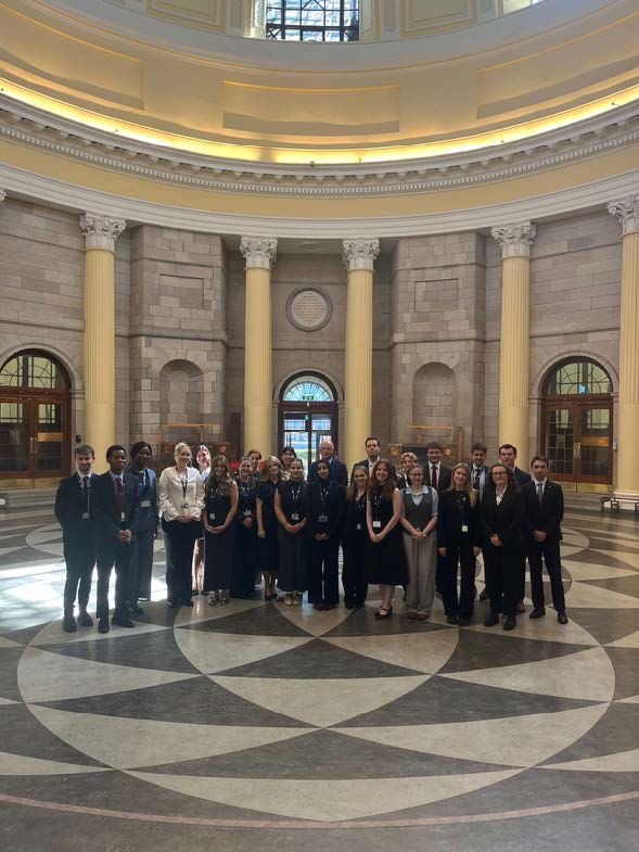 A large group of approximately 25 young adults, dressed in formal attire (suits, dresses, business casual), stand together for a photo in a grand, circular hall. The hall features tall, classical columns, stone walls, and arched doorways with large windows. The floor has a distinctive geometric pattern in light and dark tiles. The ceiling is a domed structure. The group is diverse in appearance, and most are looking towards the camera, some smiling.