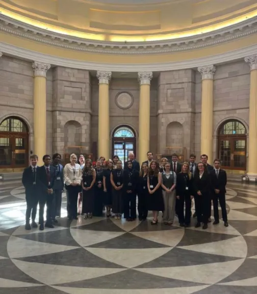 A large group of approximately 25 young adults, dressed in formal attire (suits, dresses, business casual), stand together for a photo in a grand, circular hall. The hall features tall, classical columns, stone walls, and arched doorways with large windows. The floor has a distinctive geometric pattern in light and dark tiles. The ceiling is a domed structure. The group is diverse in appearance, and most are looking towards the camera, some smiling.