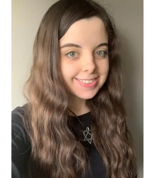 A young woman with long, wavy brown hair and light-colored eyes smiles warmly at the camera. She is wearing a black top and a necklace with a star-shaped pendant. Her hair frames her face, and she has a fresh, natural appearance. The background is a plain light grey wall.