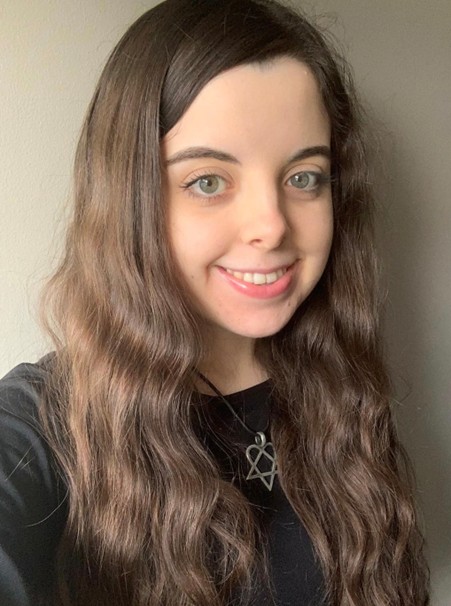 A young woman with long, wavy brown hair and light-colored eyes smiles warmly at the camera. She is wearing a black top and a necklace with a star-shaped pendant. Her hair frames her face, and she has a fresh, natural appearance. The background is a plain light grey wall.