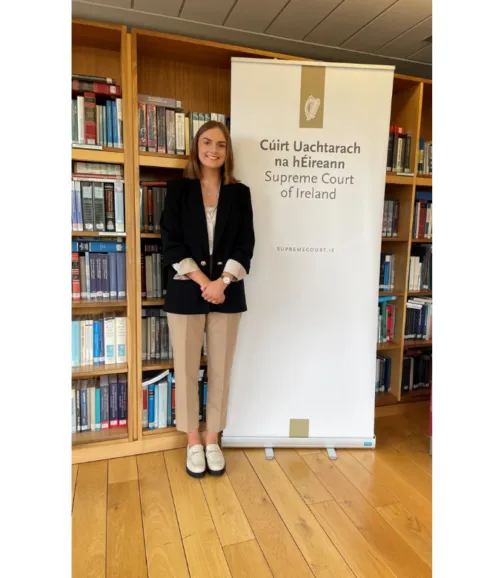 A young woman with short, straight brown hair and a bright smile stands in a library or office setting. She is wearing a black blazer over a white top, tan trousers, and white platform loafers. Her hands are clasped in front of her. To her right is a white pull-up banner with the logo and name of 