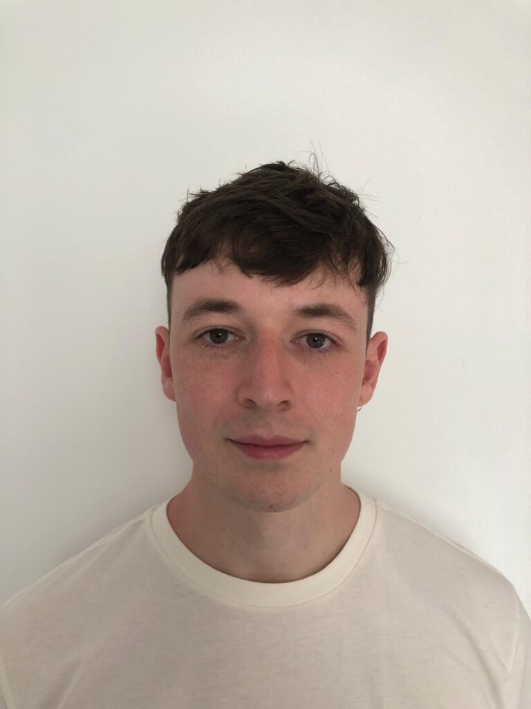 A young man with short, textured dark brown hair and light brown eyes looks directly at the camera with a neutral expression. He has fair skin and a light stubble. He is wearing a plain light-colored t-shirt. The background is a plain white wall.