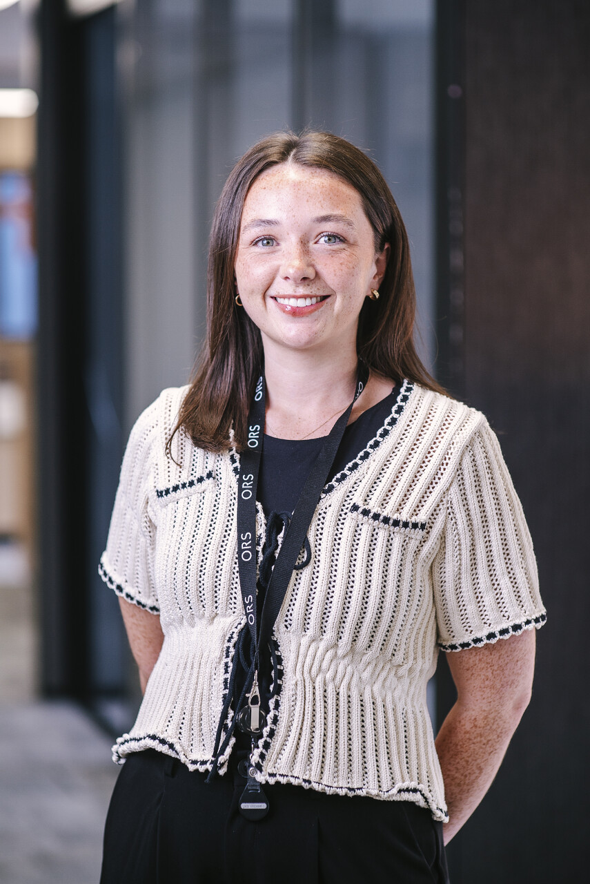 A young woman with long, dark brown hair and freckles smiles warmly at the camera. She is wearing a cream-colored, short-sleeved knit cardigan with dark trim over a black top. A black lanyard with white text that says "ORS" is hanging around her neck. Her hands are clasped behind her back. The background is a blurred indoor setting, possibly an office or modern building, with hints of blue and dark tones.