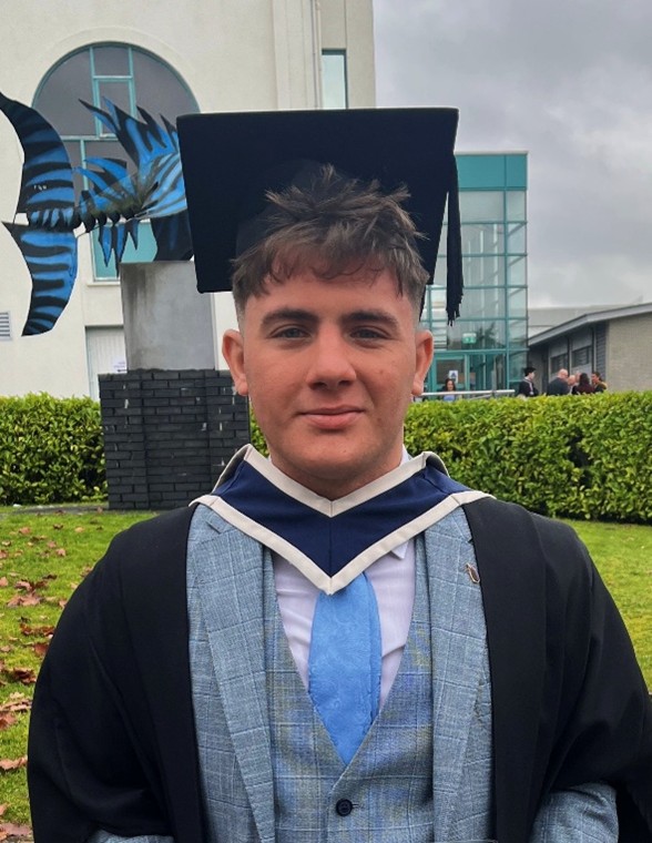 A young man with short brown hair, wearing a graduation cap, gown, and a blue and white hood, looks directly at the camera with a slight smile. Underneath his academic regalia, he is wearing a light blue plaid suit jacket, a white shirt, and a light blue tie. The background shows a modern building with a large blue sculpture and green grass, suggesting a university campus setting on an overcast day.