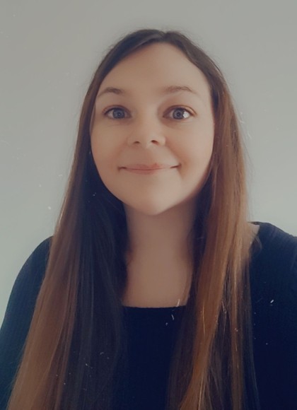 A young woman with long, straight brown hair parted in the middle, smiles gently at the camera. She has wide, light-colored eyes and is wearing a dark top. The background is a plain light-colored wall.